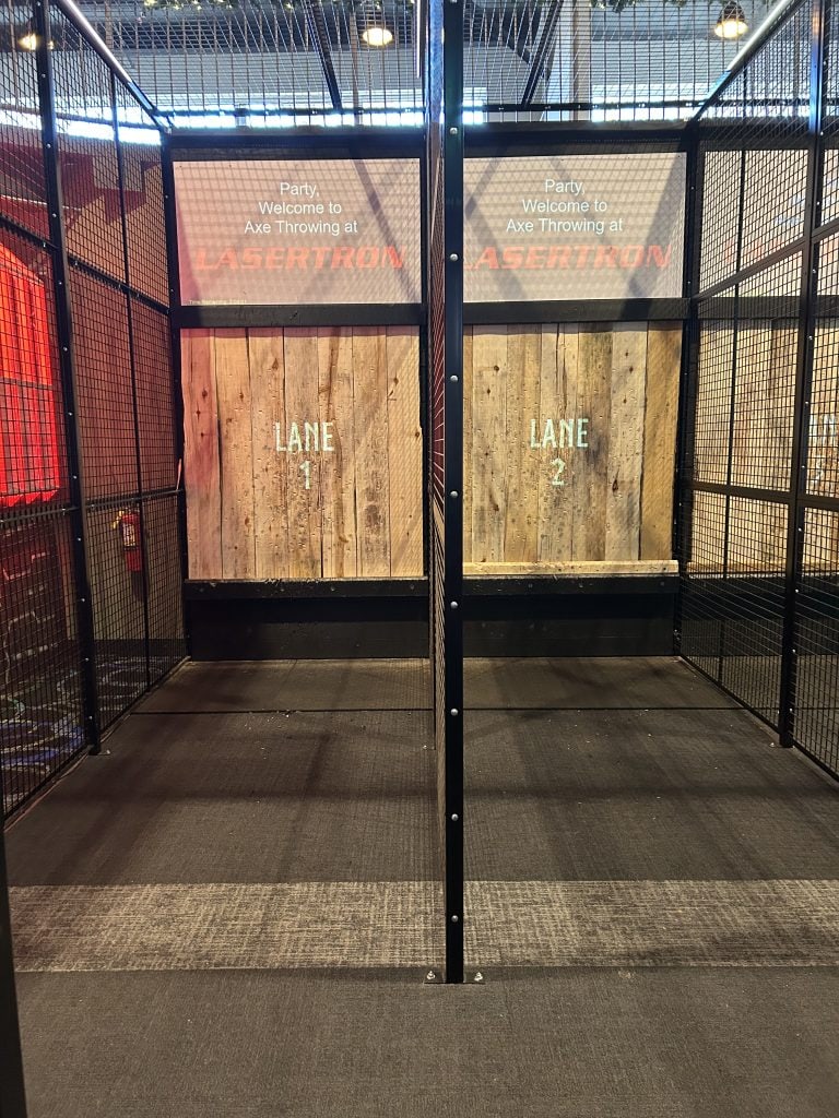 Two enclosed axe-throwing lanes with wooden targets labeled “Lane 1” and “Lane 2” sit inside a black metal cage structure. Overhead screens display a welcome message for axe throwing at LASERTRON.
