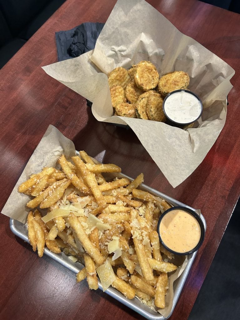 A tray of golden French fries topped with shredded Parmesan cheese is served with a side of creamy dipping sauce. Next to it, a basket of crispy fried pickle chips comes with a cup of ranch dressing.