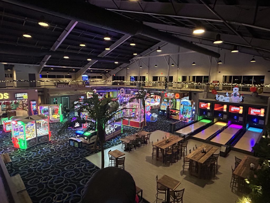Indoor arcade and entertainment space featuring colorful game machines, claw machines, and a row of four mini-bowling lanes with glowing lanes. Wooden tables and chairs are arranged in the foreground, with festive holiday decorations and string lights along the balcony.