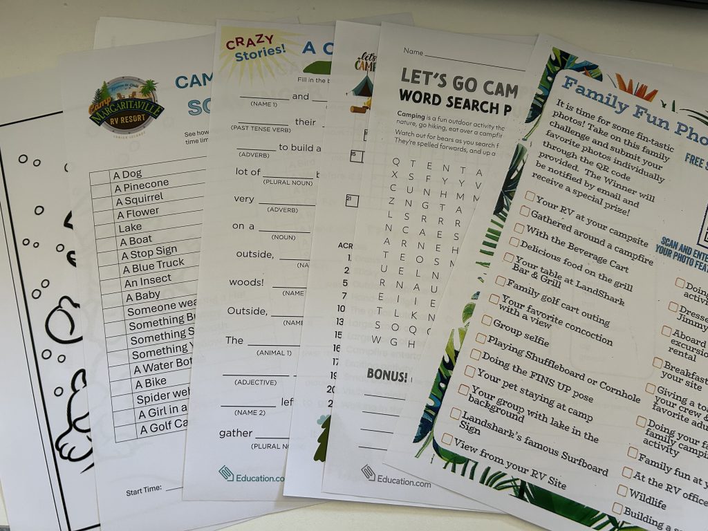 A collection of printed family activity sheets from Camp Margaritaville, including a scavenger hunt list, a “Crazy Stories” fill-in-the-blank game, a camping-themed word search, and a family fun photo challenge checklist. Bright colors and playful designs invite participation in camping and resort activities.