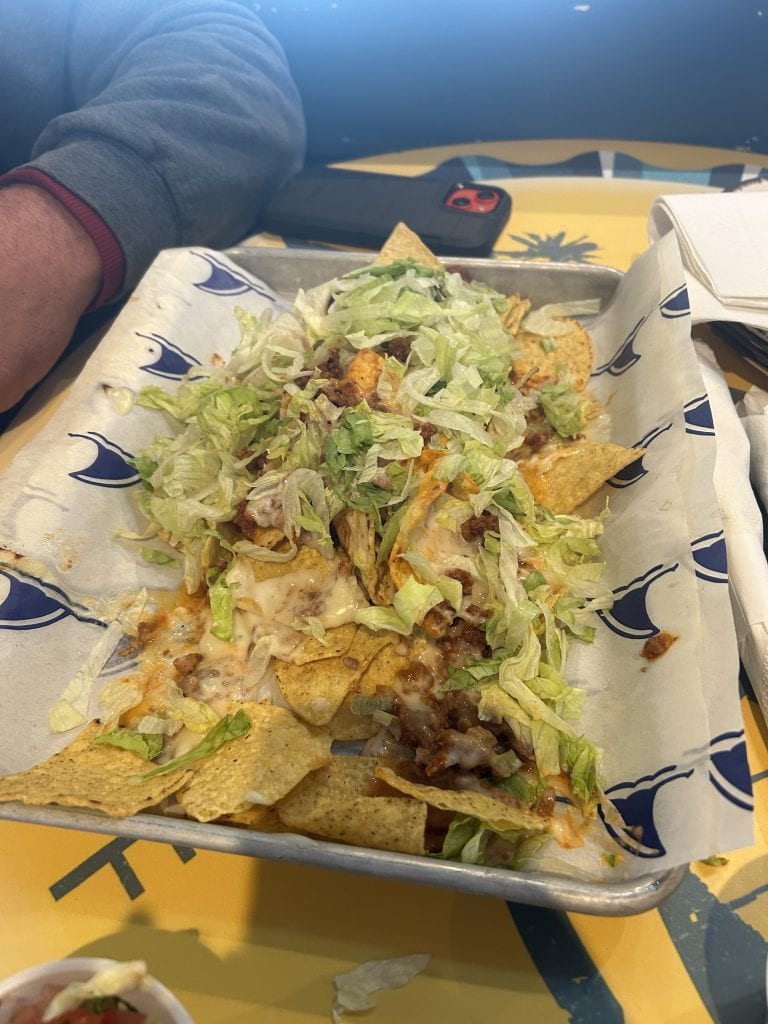 A tray of loaded nachos topped with melted cheese, seasoned ground beef, and shredded lettuce sits on branded paper. The meal rests on a yellow table with a small cup of salsa nearby.