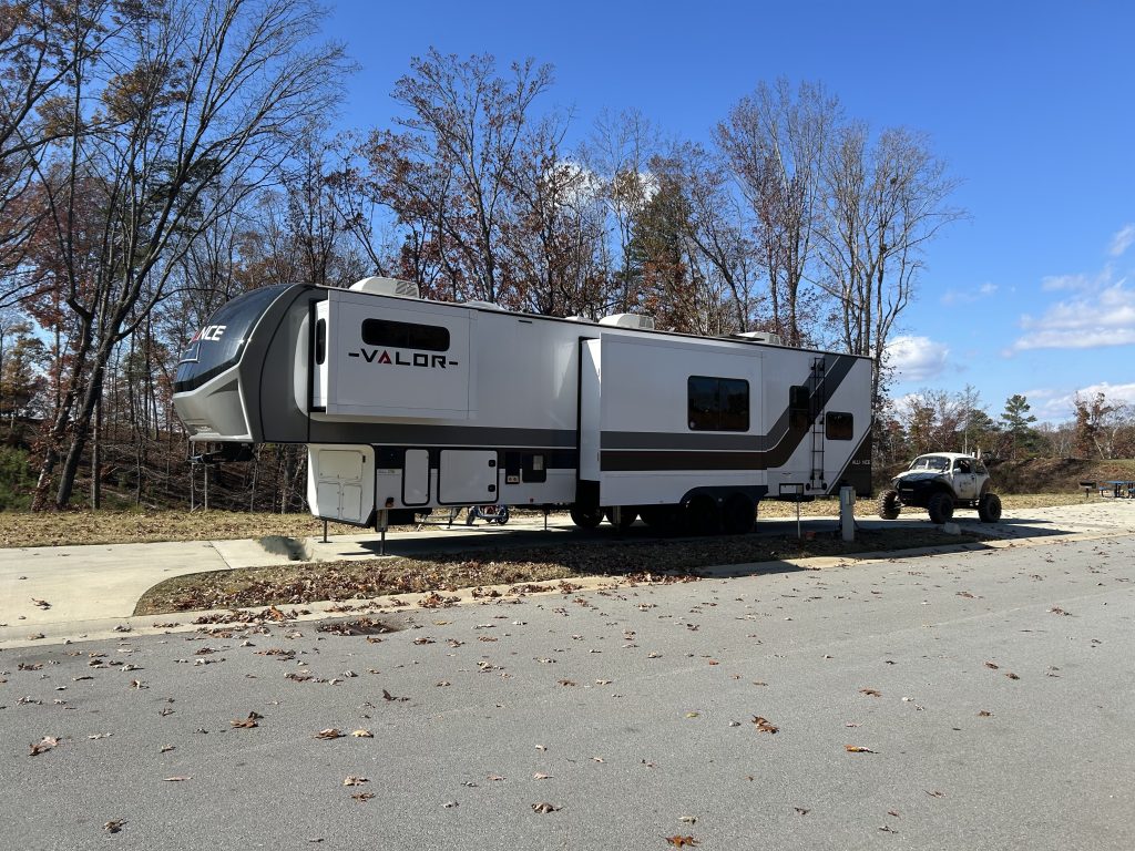 A large white and gray Valor fifth-wheel RV is parked at a paved campsite with a white off-road vehicle hitched behind. Autumn trees with sparse leaves stand in the background under a bright blue sky.