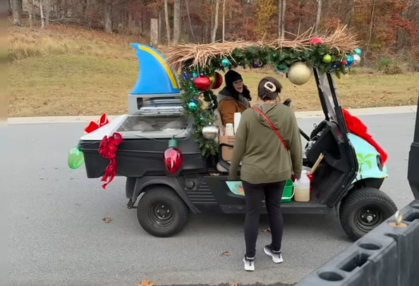 A golf cart decorated with holiday ornaments, garland, and a thatched roof serves as a mobile bar. A woman stands beside it ordering drinks from the driver, with autumn trees in the background.