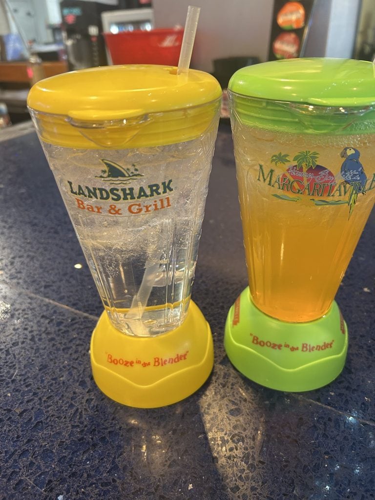 Two colorful souvenir cups from Landshark Bar & Grill and Margaritaville sit on a bar counter, each with a matching lid and base reading “Booze in the Blender.” One cup holds a clear drink with ice, and the other contains an orange beverage.