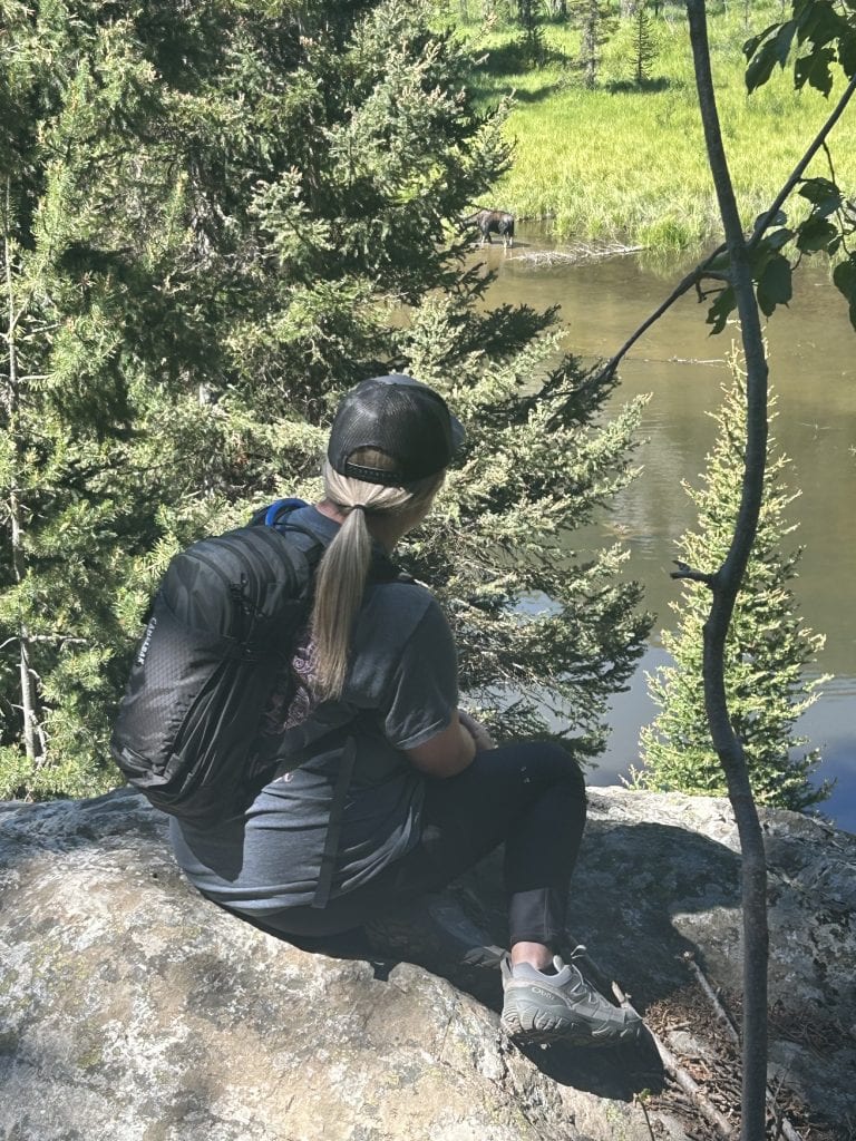 Morgan, a woman with blonde hair, is wearing a black cap, gray shirt, black leggings, and hiking shoes sits on a rock ledge with a black backpack. She looks out over a pond partially hidden by evergreen trees, where a moose stands in the shallow water near the grassy shore.