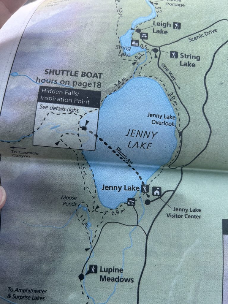 A close-up of a trail map highlights Jenny Lake in Grand Teton National Park, showing surrounding points of interest such as Lupine Meadows, Moose Ponds, Hidden Falls, Inspiration Point, String Lake, and Leigh Lake. Hiking trails, shuttle boat routes, and scenic drives are marked with distances and icons.