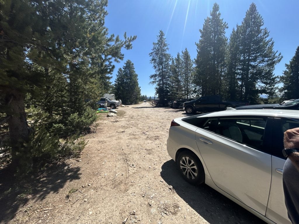 A dirt parking area surrounded by tall pine trees, with several cars and trucks parked in the sunlight. A silver sedan is in the foreground, and camping or outdoor gear is visible near a white truck in the background.