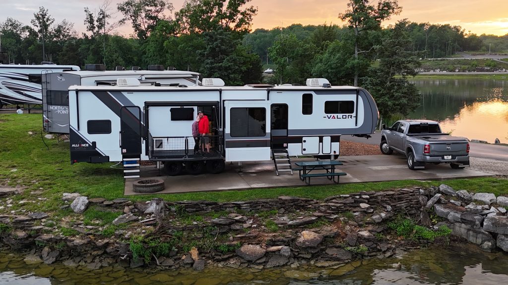 A large Alliance Valor fifth-wheel RV is parked beside a lake at sunset with a gray dually pickup truck parked in front. Alan and Morgan stand smiling on the RV’s side patio deck while surrounded by trees and water, capturing the relaxed vibe of RV travel featured on their blog - Middletons On The Move.