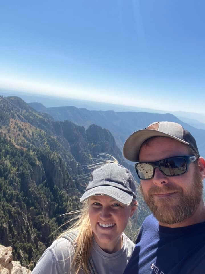 Morgan and Alan Middleton smile for a selfie at the edge of Sandia Peak, with dramatic mountain cliffs and a hazy blue horizon stretching behind them.
