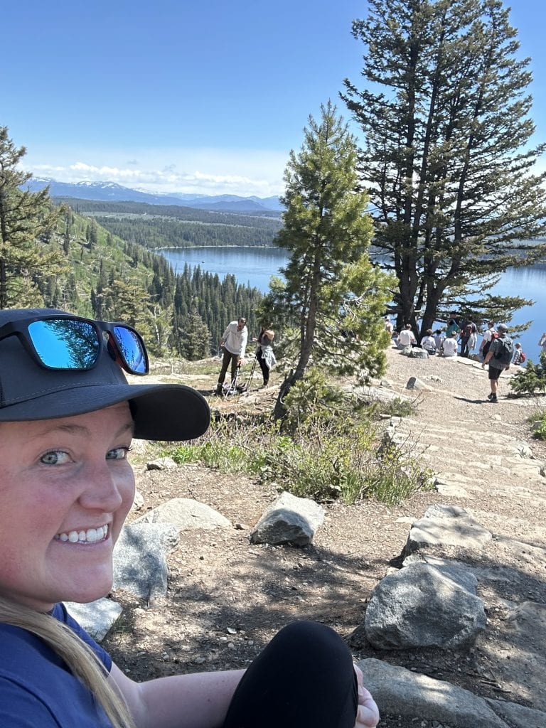 Morgan, a smiling female hiker, sits along the rocky trail to Inspiration Point, with Jenny Lake and distant mountains framed by tall pine trees in the background. The photo captures a scenic stop on the Jenny Lake hike to Inspiration Point on a clear, sunny day.