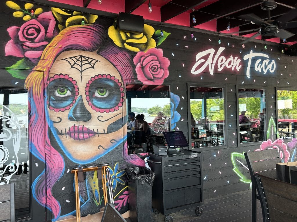 Vibrant mural on the exterior of Neon Taco, featuring a Day of the Dead-style woman with pink and purple hair, green eyes, and decorative face paint, surrounded by colorful roses. The restaurant’s name is painted in bold white script above the artwork, with string lights and patio seating visible.
