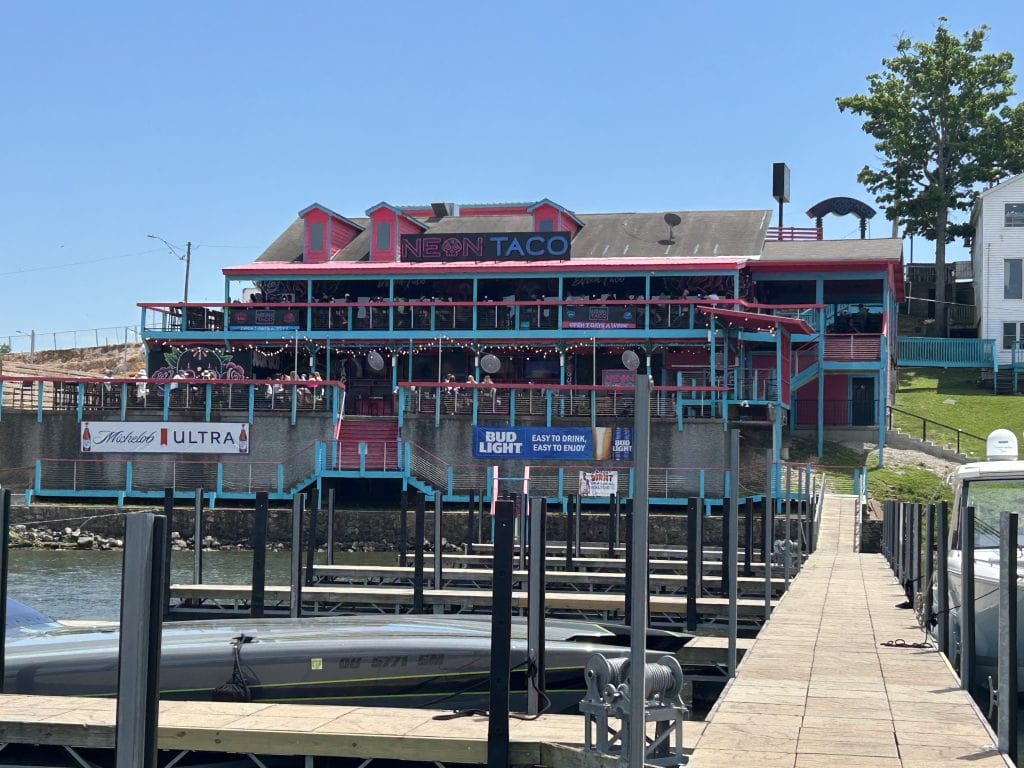 Exterior view of Neon Taco, a vibrant pink and turquoise waterfront restaurant at Lake of the Ozarks. The multilevel building features large outdoor dining decks lined with string lights, a staircase leading up from a boat dock, and branded banners for Michelob Ultra and Bud Light on the lower level.