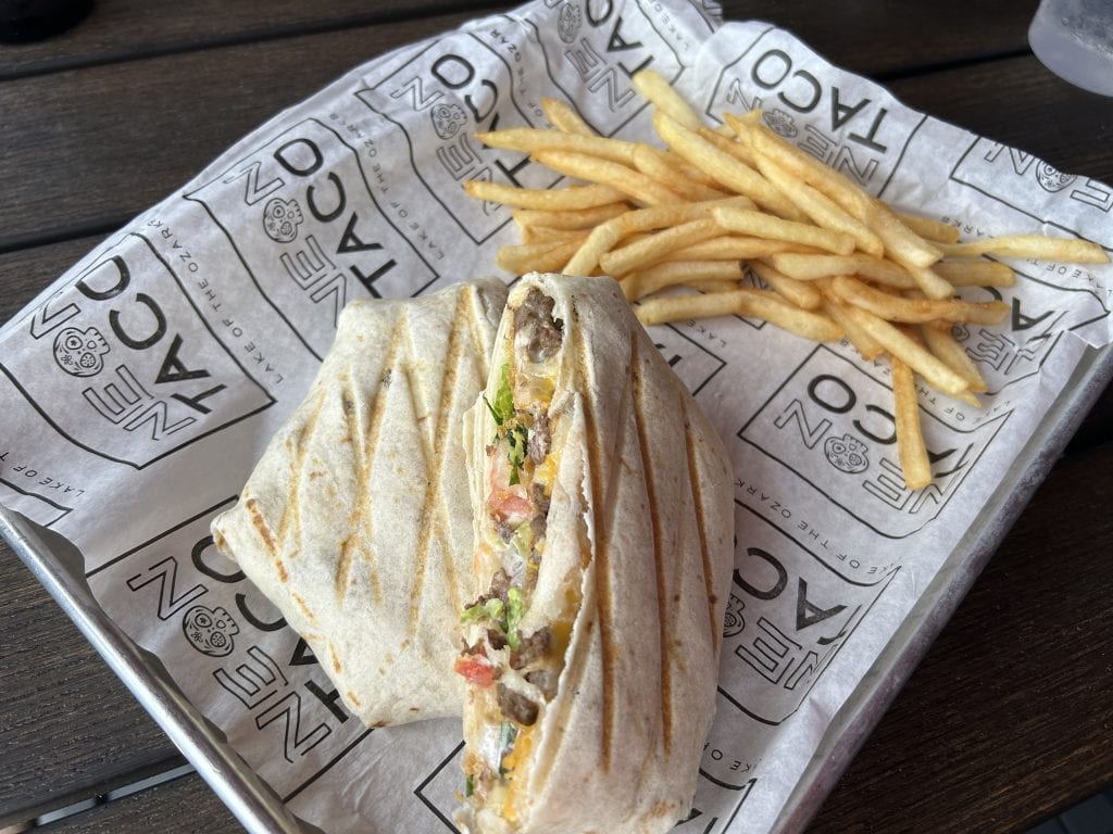 A grilled crunch wrap filled with seasoned beef, melted cheese, lettuce, and tomato, served with a side of golden French fries on branded Neon Taco paper at Lake of the Ozarks. The food is presented on a metal tray atop a dark wooden table.