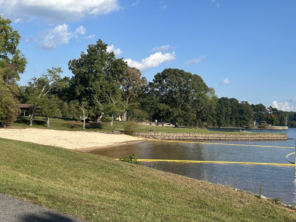 A roped-off sandy swim area at Old Federal Campground curves along the edge of Lake Lanier, bordered by grassy banks and stone retaining walls. Trees and picnic shelters are visible in the background on a sunny afternoon.