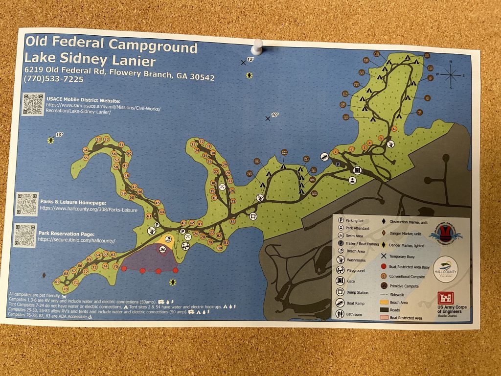 A detailed map of Old Federal Campground at Lake Sidney Lanier is pinned to a corkboard, showing campsite numbers, roads, swim areas, boat ramps, and amenities like bathrooms and playgrounds. The map includes QR codes for the park's website and reservation page, along with a legend explaining symbols for different campsite types and park features.