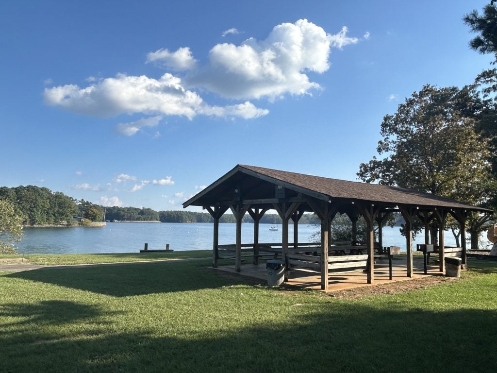 A wooden picnic pavilion at Old Federal Campground sits near the edge of Lake Lanier, surrounded by green grass and shaded by trees. Calm water and anchored boats can be seen in the distance under a bright blue sky with scattered clouds.