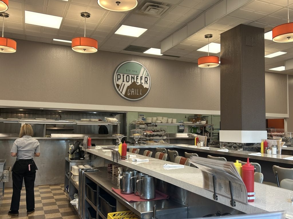 The interior of Pioneer Grill features a long retro-style counter lined with stools, condiment bottles, and menus. A waitress stands near the open kitchen, where shelves hold plates and baked goods, beneath a large circular Pioneer Grill sign on the wall.