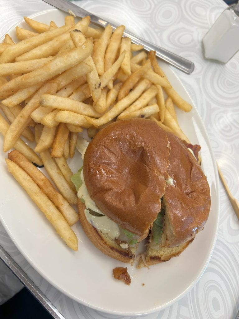 A white plate holds a sandwich on a toasted bun with lettuce and sauce, served alongside a generous portion of golden French fries. The sandwich is cut in half, revealing its fresh ingredients inside.