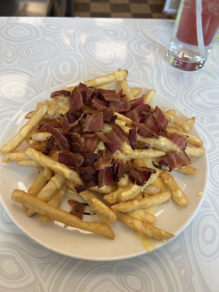 A plate of golden French fries is topped with melted cheese and generous pieces of crispy bacon. In the background, a partially filled glass of pink beverage with a straw sits on the counter.