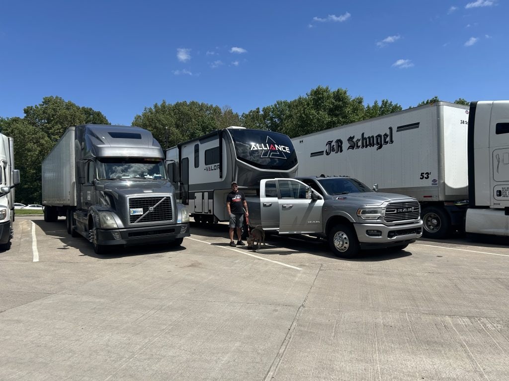 A silver Ram dually towing an Alliance Valor fifth-wheel RV is parked at a rest area alongside semi-trucks during a road trip, with a man and dog standing beside the truck under a clear blue sky.