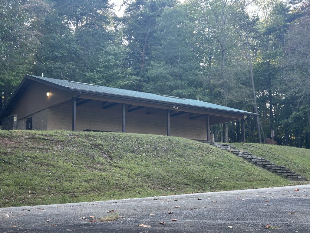 Restroom and shower facility at River Forks Park on Lake Lanier, built into a grassy hillside with a green metal roof and a covered porch supported by beams. A set of stone steps leads up from the paved road to the building, surrounded by dense forest.