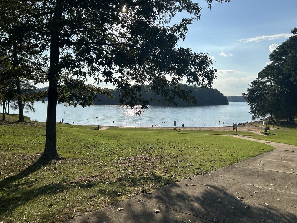 View of the sandy swimming beach at River Forks Park on Lake Lanier, with people wading and playing in the water. A paved walkway leads down through a grassy area shaded by trees, with calm lake water and forested shoreline in the background.