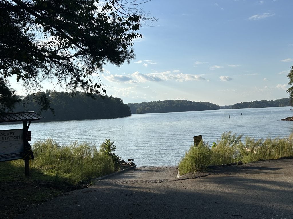 Scenic boat ramp at River Forks Park on Lake Lanier, leading directly into calm, blue water surrounded by lush green shoreline and distant forested islands. A life jacket station is partially visible on the left, with tall grass framing the ramp under a clear sky.