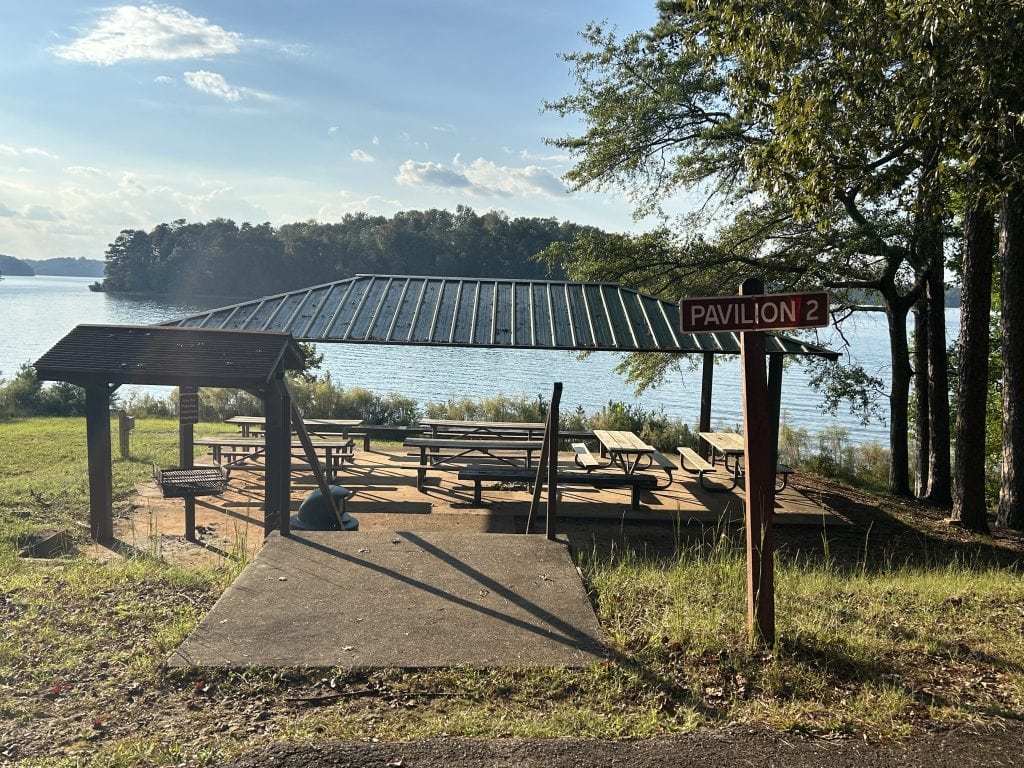 Pavilion 2 at River Forks Park on Lake Lanier, a covered lakeside picnic area with multiple tables, a grill, and scenic water views. The metal-roofed shelter sits on a concrete pad surrounded by trees and grassy shoreline under a clear sky.