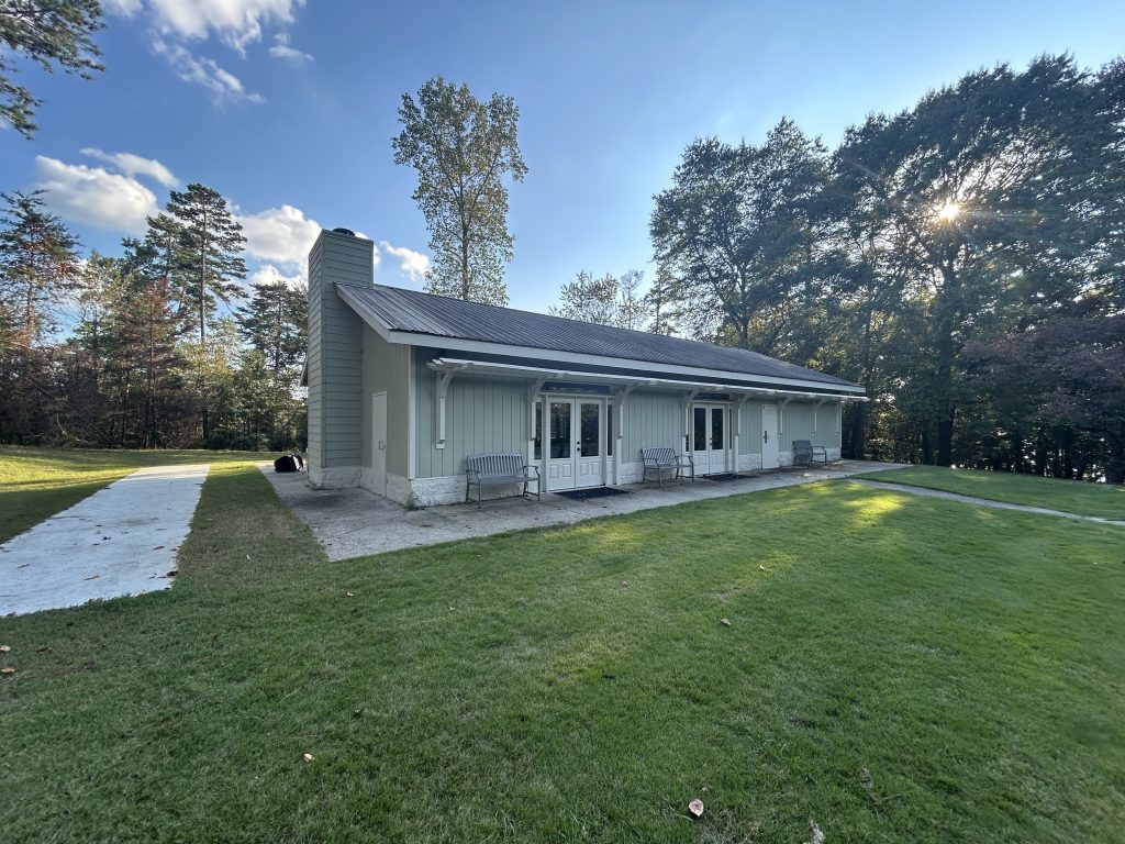 The Retreat at River Forks Park, a light gray event and conference building with a metal roof, large windows, and multiple benches on a concrete patio. Surrounded by green lawn and tall trees, the space is available for rent for gatherings, meetings, and special events.