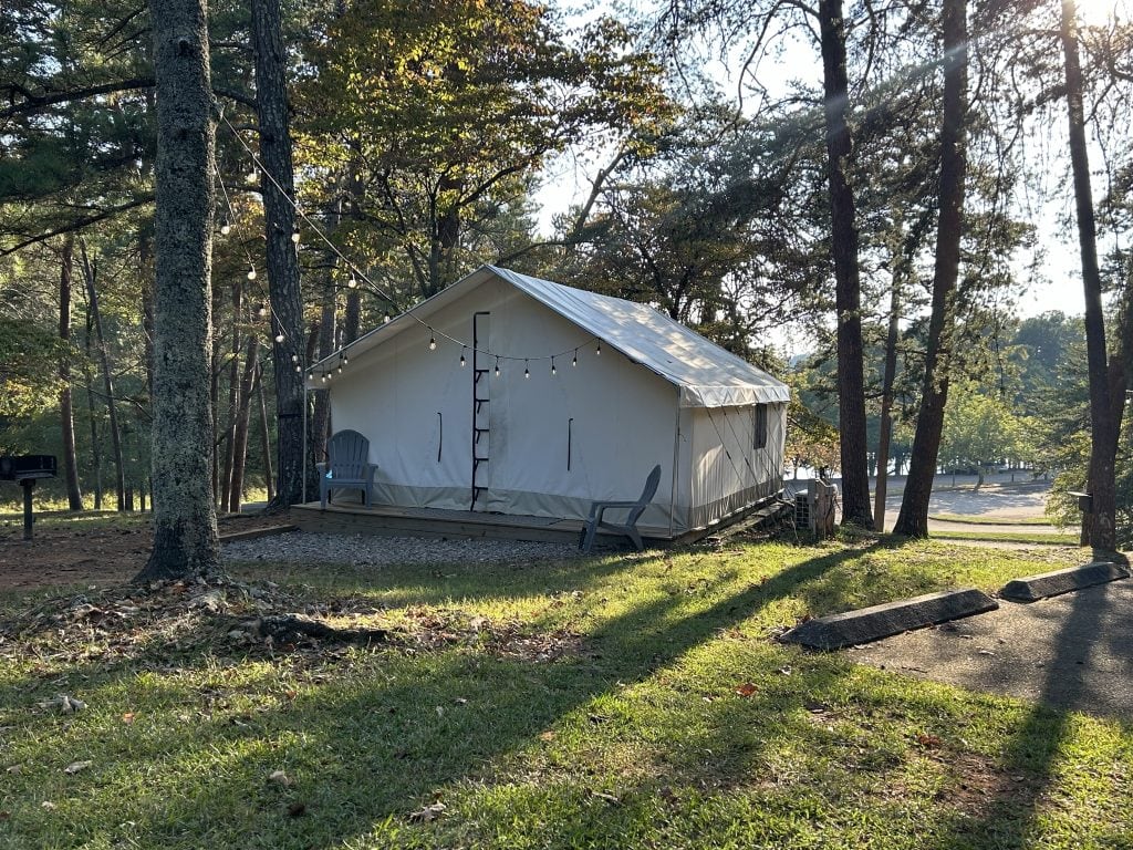Canvas glamping tent at River Forks Park on Lake Lanier, set on a raised wooden platform with string lights, two Adirondack chairs, and a wooded lake view in the background. The site includes a nearby grill and parking space, surrounded by tall trees and soft evening light.