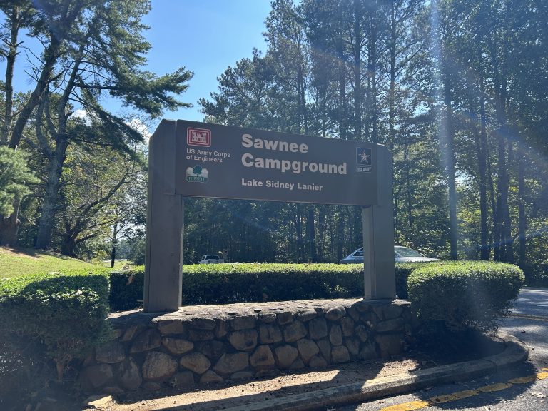 Brown and gray entrance sign for Sawnee Campground on Lake Lanier, maintained by the U.S. Army Corps of Engineers, surrounded by trimmed hedges and tall pine trees on a sunny day. A white car is partially visible behind the sign along the curved campground road.