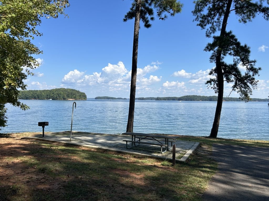 A shaded lakeside picnic area with a concrete pad, picnic table, grill, and tall shower pole overlooks calm blue water. In the distance, tree-covered islands dot the horizon beneath a bright blue sky with scattered clouds.