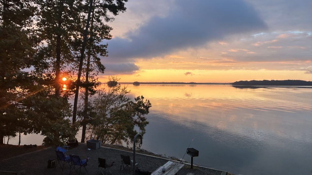 Golden sunrise over a calm lake, with the sun peeking through tall trees and reflecting on the water’s surface. In the foreground, camping chairs, a picnic table, and a grill sit along the shoreline, adding a peaceful, outdoor atmosphere.
