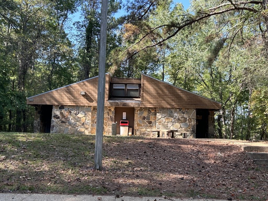 Stone and wood restroom building at Shoal Creek Campground surrounded by trees, with a sloped shingle roof and a red trash bin outside the entrance. The facility sits on a small hill with a bench and stair access from the paved path below.