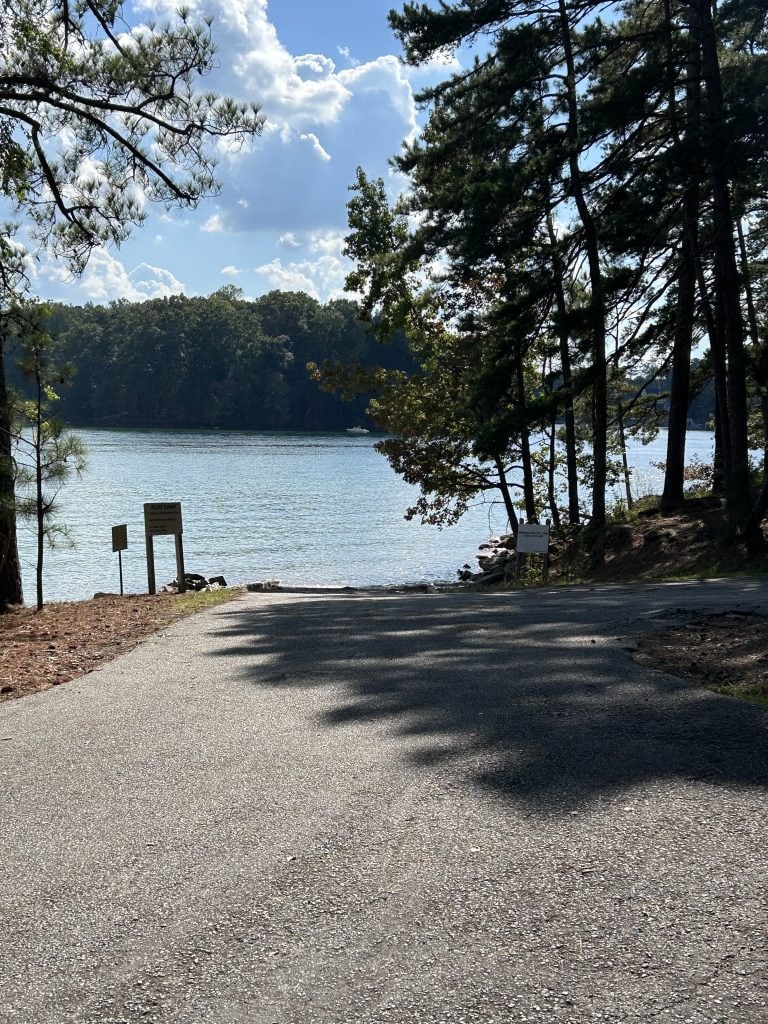 Paved boat ramp at Shoal Creek Campground leading into Lake Lanier, surrounded by tall pine trees and partially shaded by their branches. Signs are posted near the water’s edge, with calm lake water and a forested shoreline visible in the background under a partly cloudy sky.