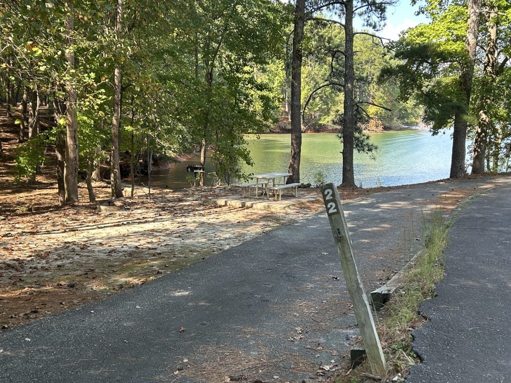 Campsite 22 at Shoal Creek Campground with a paved driveway, picnic table, and grill set in a wooded area beside a quiet cove. The site is surrounded by tall trees and overlooks calm, green-tinted water with scattered autumn leaves on the ground.