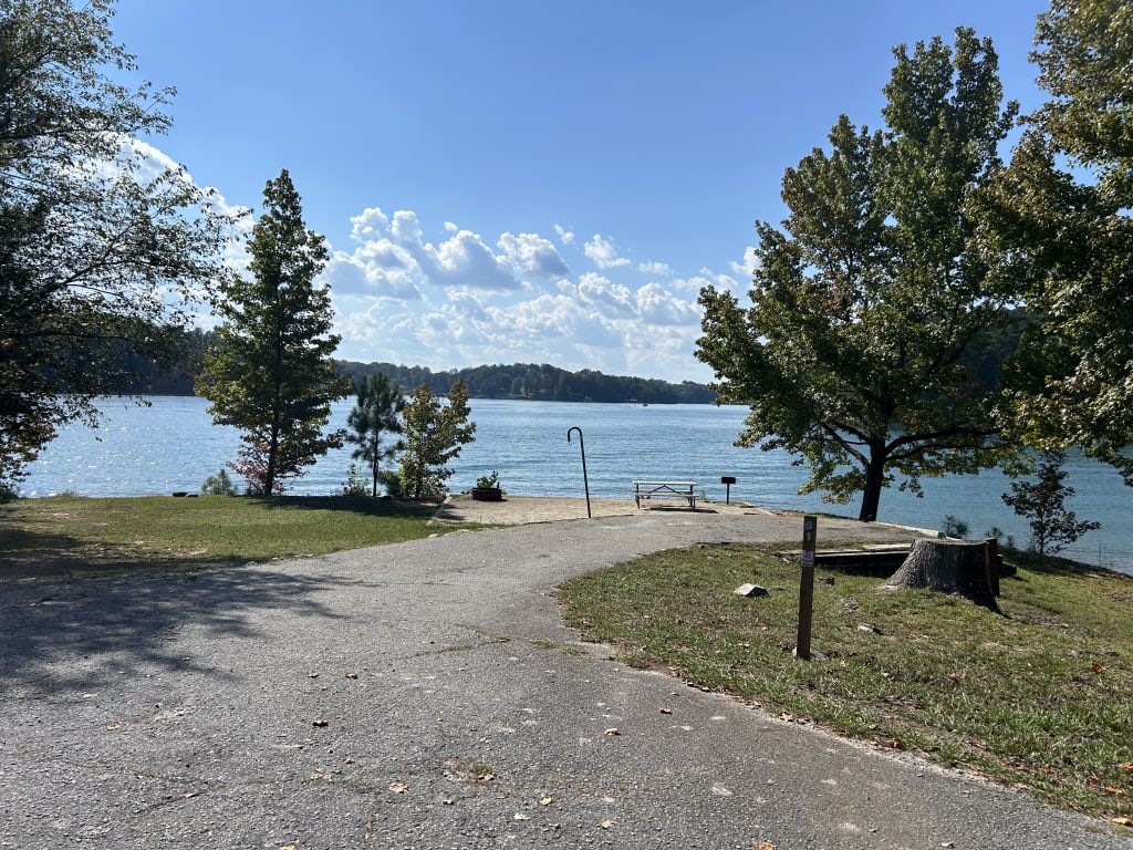 Lakefront campsite at Shoal Creek Campground with a concrete pad, picnic table, grill, and water spigot overlooking blue water and wooded hills in the distance. The site is partially shaded with a tree stump and fire ring near the paved drive, surrounded by scattered fall leaves.