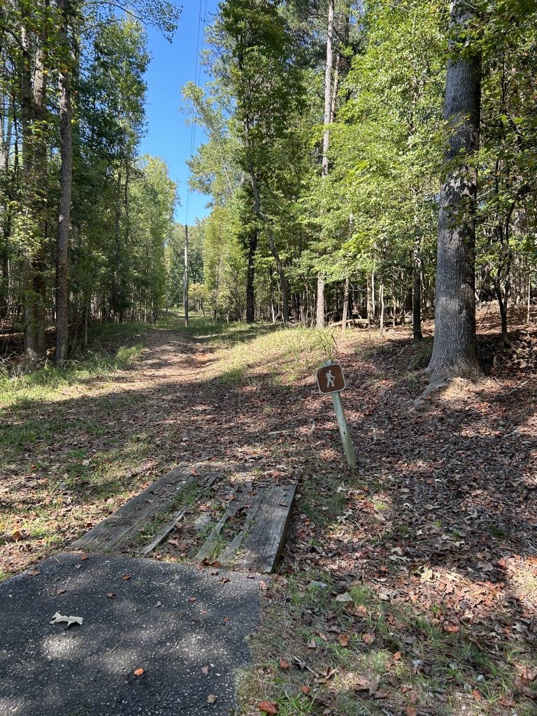Trailhead at Shoal Creek Campground marked by a brown hiker sign beside a wooden footbridge leading into a wooded path under power lines. Fallen leaves cover the ground, and the trail stretches into a sunlit forest surrounded by tall trees.