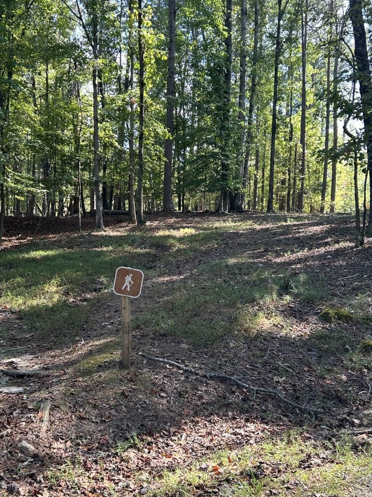 Wooded hiking trail entrance at Shoal Creek Campground marked by a small brown sign with a hiker icon. The trail begins on a gently sloping forest floor surrounded by tall trees and dappled sunlight.