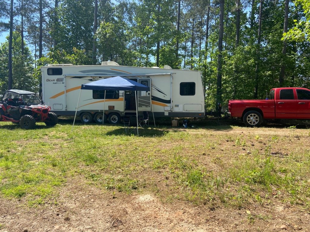 A toy hauler RV with an awning setup is parked at a wooded campsite, flanked by a red pickup truck and a red side-by-side off-road vehicle under sunny skies.