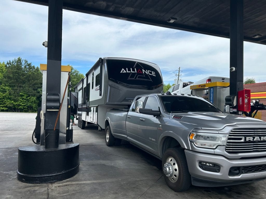 A silver Ram heavy-duty pickup truck towing a large Alliance fifth-wheel RV is parked at a gas station pump under a covered canopy. The scene shows the truck refueling while traveling, with trees and other fuel pumps in the background.
