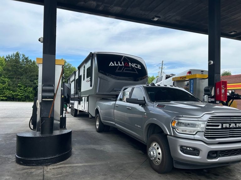 A silver Ram heavy-duty pickup truck towing a large Alliance fifth-wheel RV is parked at a gas station pump under a covered canopy. The scene shows the truck refueling while traveling, with trees and other fuel pumps in the background.