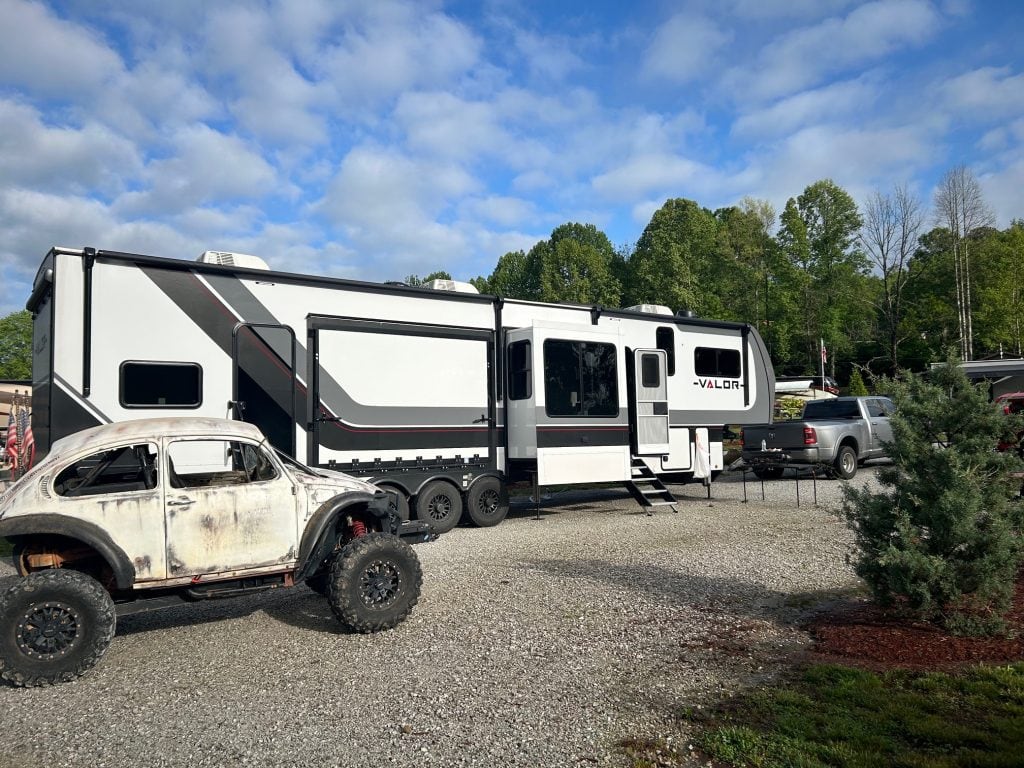 A white Alliance Valor fifth-wheel RV is parked in a gravel lot, hitched to a silver pickup truck. In the foreground, a weathered Volkswagen Beetle body is mounted on a lifted side-by-side frame with oversized off-road tires, set against a backdrop of trees and other RVs under a partly cloudy sky.