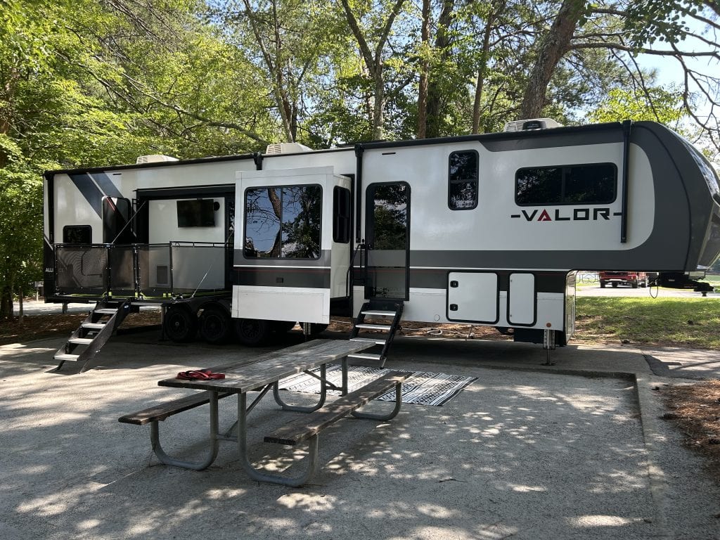 A modern Valor fifth-wheel RV is set up at a shaded campsite with slide-outs extended and a picnic table in the foreground, surrounded by tall trees on a sunny day.