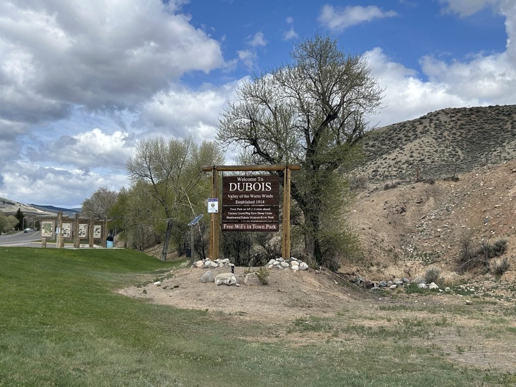 A wooden welcome sign at the edge of Dubois, Wyoming reads “Welcome to Dubois, Valley of the Warm Winds,” with additional text noting the town’s elevation and nearby destinations, set against rolling hills and a partly cloudy sky.