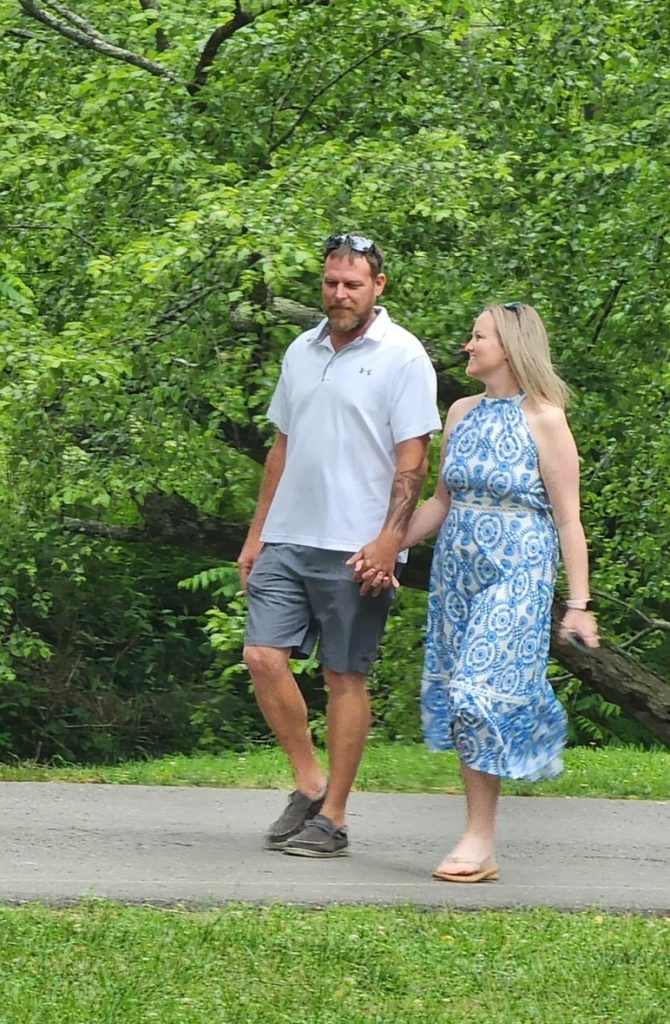 Alan and Morgan walk hand in hand along a paved path surrounded by lush green trees, enjoying a peaceful moment outdoors on a summer day.