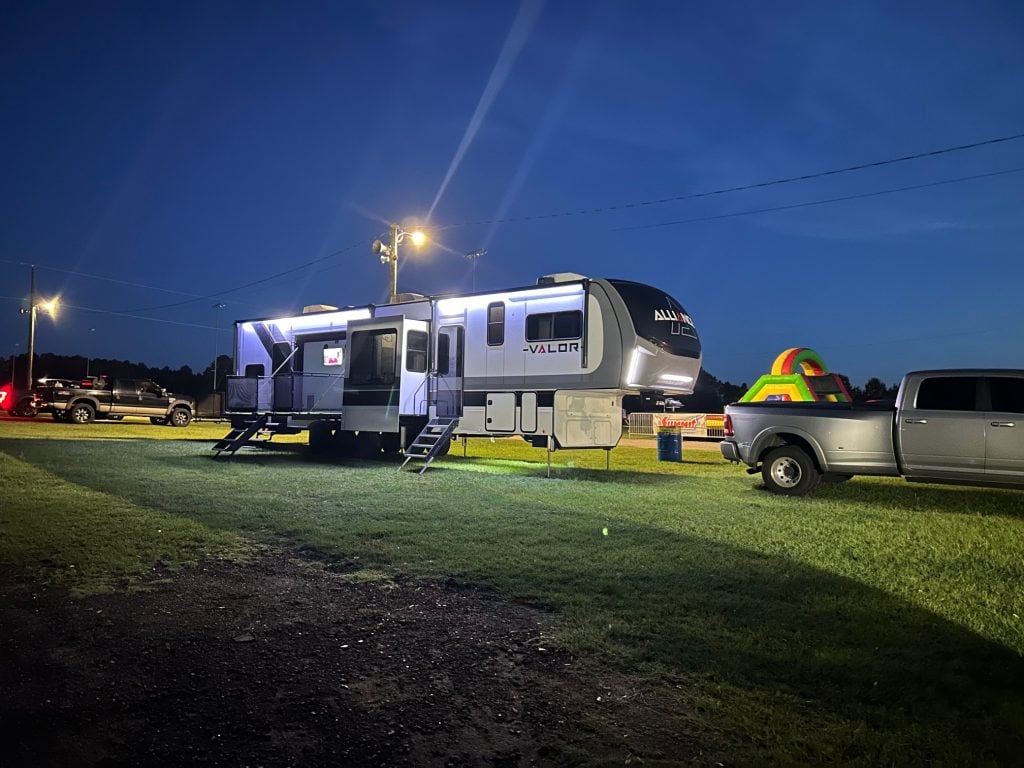A white, black, and silver Alliance Valor fifth-wheel RV glows under bright exterior lights at night. In front of it is a silver dually pickup truck on a grassy lot. In the background, a colorful inflatable play structure and other vehicles are visible under the dark blue evening sky.