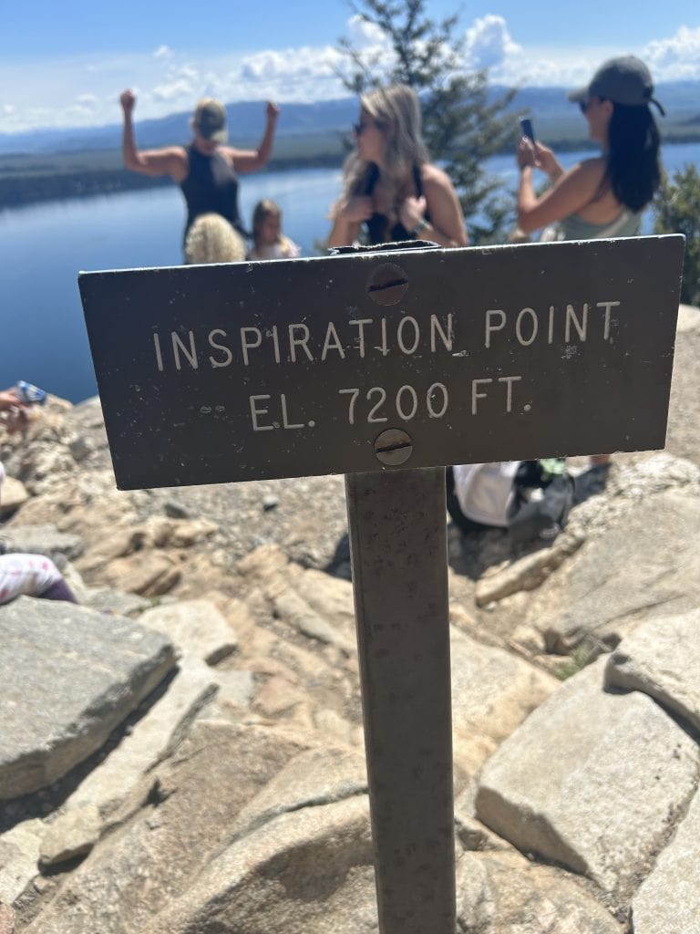 A metal sign reading “Inspiration Point EL. 7200 FT.” stands at the summit overlook, marking the end of the Jenny Lake hike to Inspiration Point. Behind it, hikers celebrate and take photos with stunning lake and mountain views in the background.