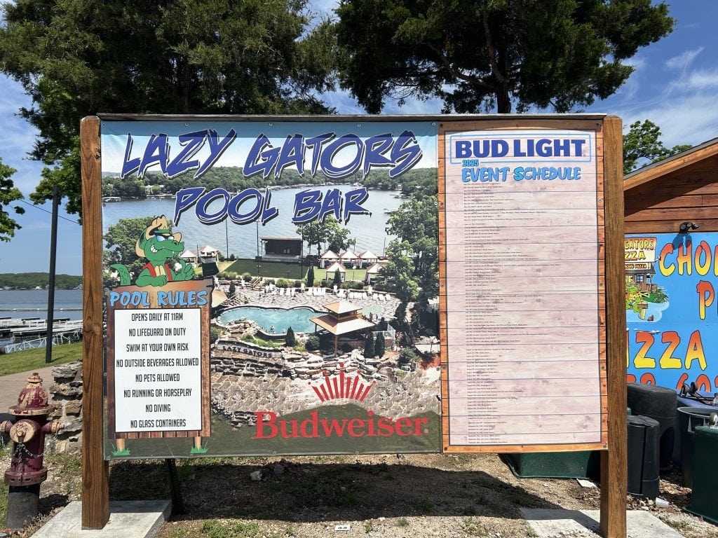 Signboard at Lazy Gators Pool Bar at Lake of the Ozarks, Missouri, featuring pool rules and a Bud Light-sponsored 2025 event schedule. The board includes a cartoon alligator and a photo of the pool area with cabanas and the lake in the background. Bright, sunny day with trees and lake visible around the sign.