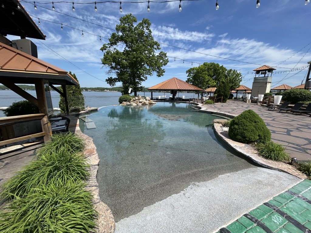 Infinity-edge pool at Lazy Gators in Missouri overlooking the Lake of the Ozarks, with stone walkways, cabanas with copper roofs, and string lights overhead. Lush landscaping surrounds the pool, and the lake stretches out in the background under a blue sky with wispy clouds.